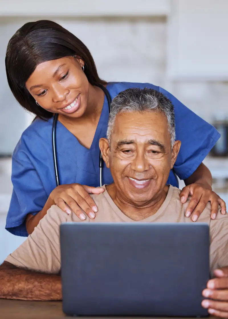 a younger caretaker standing behind an older man with him working on his laptop