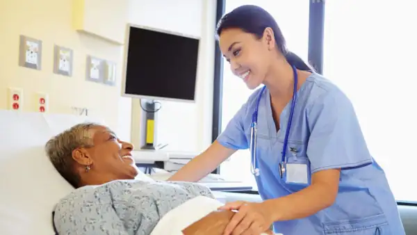a younger caretaker talking with an older woman laying in a hospital type bed