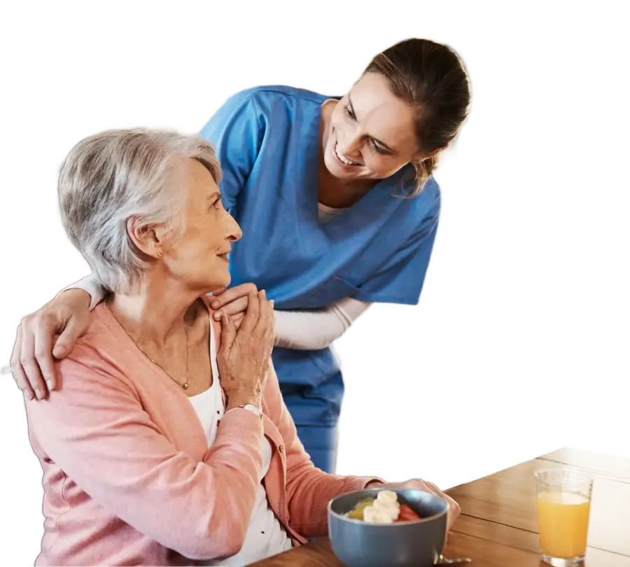 a younger caretaker with her hands on an older woman's shoulders talking with her before she eats