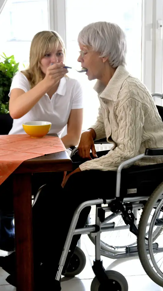 a younger caretaker feeding an older woman in a wheel chair