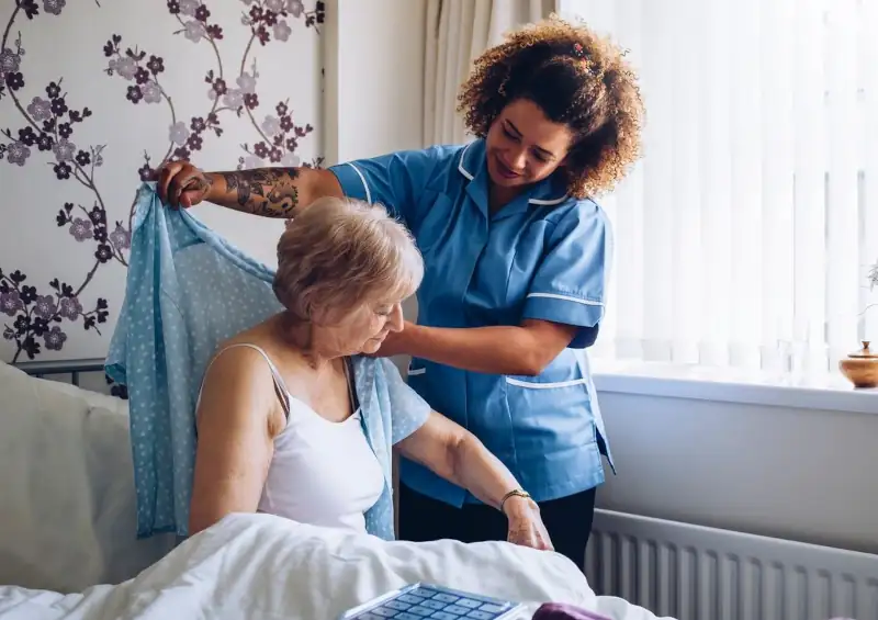 a younger caretaker helping an older woman get dressed