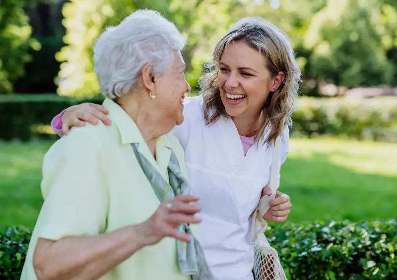 a younger caretaker walking and smiling with an older woman while they walk outside