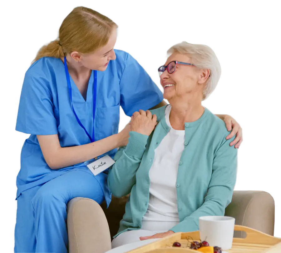 a younger caretaker sitting on the arm of a chair while talking an older woman before she eats her meal
